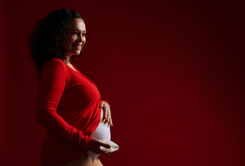 Delightful pregnant woman dressed in red shirt, holding a white orchid flower, smiling cutely,...