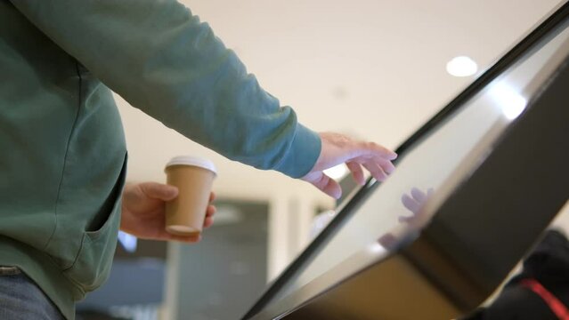 Young male hands dressed in jacket swiping for information in kiosk on background of lights of shopping mall. No face man with interactive wayfinding kiosk