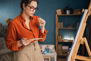 side view focused ethnic woman in casual clothes stands near the canvas holds a palette with paints...