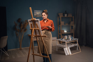 portrait of a beautiful female artist, stands alone in a modern workshop, paints a picture on canvas with paints, looks absorbed, enjoys a creative hobby for the soul. Workshop or art clas