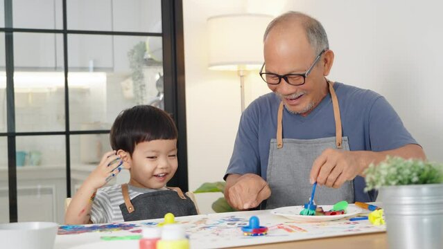 Cheerful Asian Japanese Senior man and little child boy in apron Enjoying painting art at home together. Happy Grandfather and Grandchild playing and laughing. Positive emotion.	