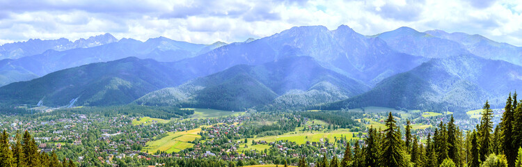 Fototapeta na wymiar Panorama of the Tatra Mountains (Poland) with the Giewont mountain seen from Gubałówka on a sunny summer day with the sun breaking through the clouds. In the foreground, below, the city of Zakopane.