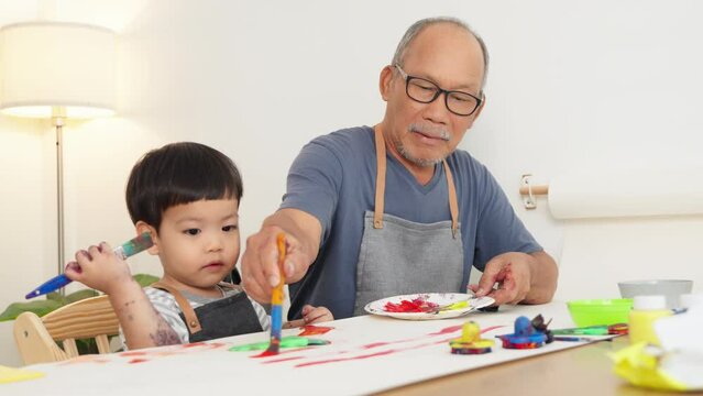 Asian Japanese Senior man and little child boy in apron painting art at home together. Happy Grandfather and Grandchild with watercolor.	