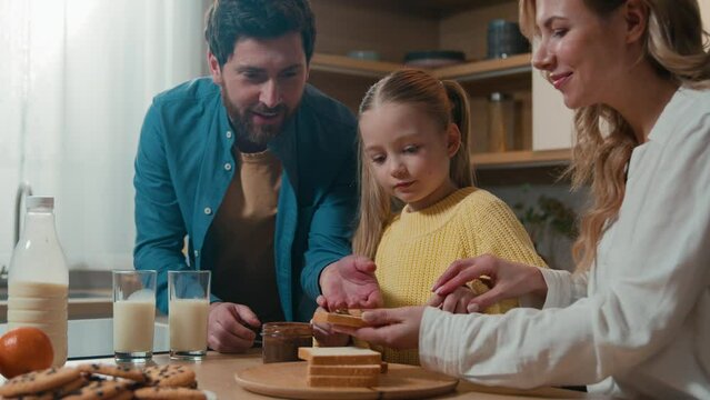 Happy Caucasian family cooking breakfast together at kitchen mother help teach little daughter child girl prepare toasts spread chocolate paste on bread father enjoy food cheerful parents and cute kid