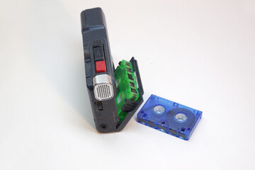 tape voice recorder, handheld, with green microcassette in cover of tape cassette compartment. Red button of Rec, silvery electret condenser mic housing, blue cassette laying near