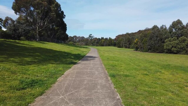Handheld walking perspective of a concrete footpath with Australian native Eucalyptus trees on sides. Public walking trail with large green outdoor area. Werribee river trail, Melbourne VIC Australia.