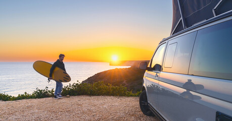 Surfer boy near his mini van and looking on the ocean at summer sunset  with a surfboard on his side