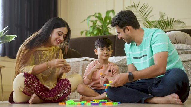 Happy indian parents playing with preteen kid with toys while sitting on floor at home - concept of family time, parenthood and