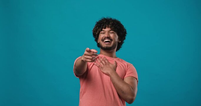Young indian man pointing finger to camera, laughing out loud, taunting making fun of ridiculous appearance, funny joke standing isolated over blue studio background