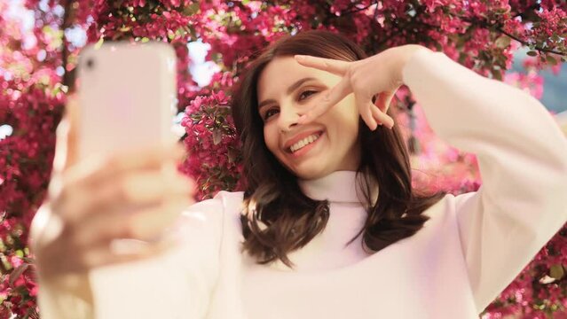 Portrait of smiling dark haired woman posing takes selfies and streams video to social media from her smartphone with pink flowers blossom on the background outdoors