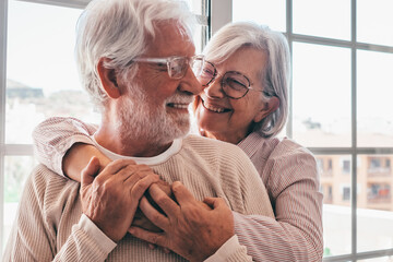 Smiling senior family couple of Caucasian people hugging in front the window, elderly man and woman with glasses embracing each other in a moment of tendress