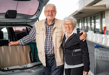 Happy lovely elderly traveler couple unload their suitcases from the parked car outside the departures area of the airport. Old senior man and woman leaving for vacation