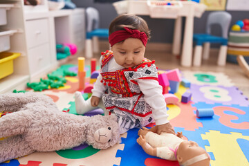 Adorable hispanic baby sitting on floor with relaxed expression at kindergarten