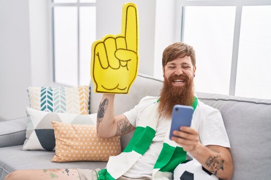 Young Redhead Man Supporting Soccer Team Using Smartphone Holding Big Hand At Home