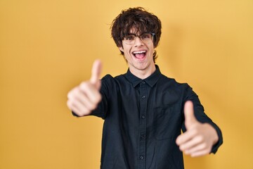 Young man wearing glasses over yellow background approving doing positive gesture with hand, thumbs up smiling and happy for success. winner gesture.