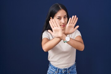 Young hispanic woman standing over blue background rejection expression crossing arms and palms doing negative sign, angry face
