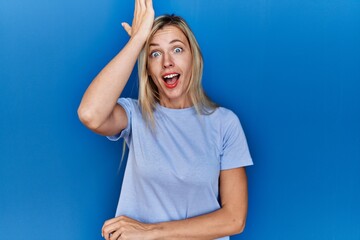 Beautiful blonde woman wearing casual t shirt over blue background surprised with hand on head for...