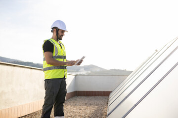 A specialized engineer in protective clothing is examining solar panels to heat the hot water in the houses using a digital tablet. Concept of solar panels, monitoring of solar panels.