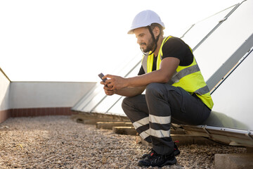 A specialized technician in protective clothing is sitting while resting and using his mobile phone. Concept of solar panels, renewable energy. Green energy.