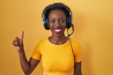 African woman with curly hair standing over yellow background wearing headphones showing and pointing up with finger number one while smiling confident and happy.