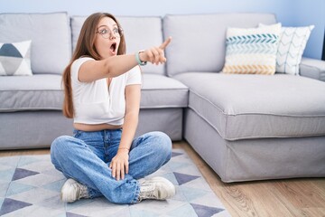 Young caucasian woman sitting on the floor at the living room pointing with finger surprised ahead, open mouth amazed expression, something on the front