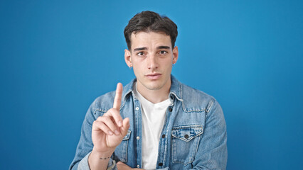 Young hispanic man standing with serious expression saying no with finger over isolated blue...
