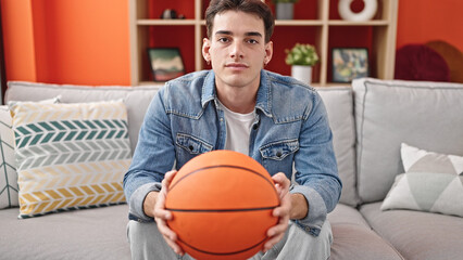 Young hispanic man holding basketball ball sitting on sofa at home