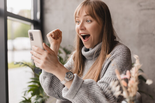 Joyful blonde woman playing video game on mobile phone, reading good news, celebrating winning lottery, feeling so happy and cheerful while making winner gesture. Wow emotions.