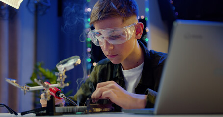 Likable smiling skillful teenage boy in protective glasses soldering small parts of panel using...