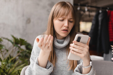 Confused puzzled blonde bang woman in casual clothes, sits in cafe, holds a smartphone in her hand, looks questioningly at the mobile phone, spreading her arm around. Girl getting surprising bad news.