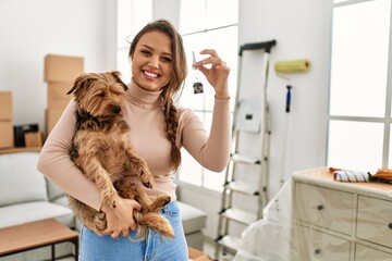 Young beautiful hispanic woman hugging dog holding key at new home