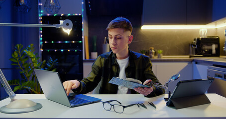 Teenage schoolboy using laptop and holding computer board in his hands and reading instructions of its structure on laptop screen. Education concept.