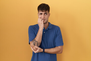 Young hispanic man standing over yellow background looking stressed and nervous with hands on mouth biting nails. anxiety problem.