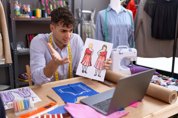 Young hispanic man tailor having video call showing clothing design at atelier