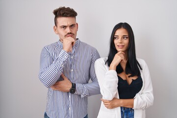 Young hispanic couple standing over white background with hand on chin thinking about question, pensive expression. smiling and thoughtful face. doubt concept.