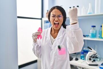Young hispanic doctor woman working at scientist laboratory holding pink ribbon annoyed and frustrated shouting with anger, yelling crazy with anger and hand raised