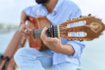Young hispanic man musician playing classical guitar sitting on balustrade at seaside