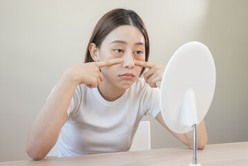 Sleepless Asian woman looking her face in the mirror and worry about dark circles under eyes