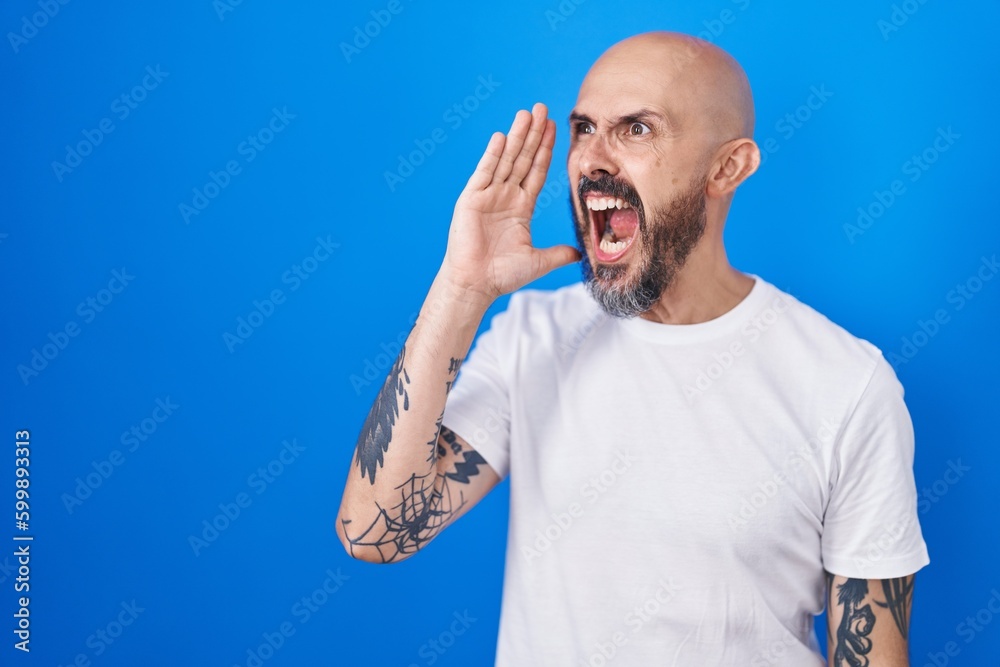 Sticker Hispanic man with tattoos standing over blue background shouting and screaming loud to side with hand on mouth. communication concept.