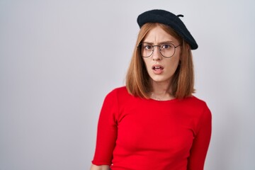 Young redhead woman standing wearing glasses and beret in shock face, looking skeptical and sarcastic, surprised with open mouth