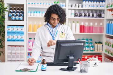 Hispanic man with curly hair working at pharmacy drugstore checking the time on wrist watch, relaxed and confident