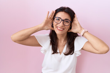 Middle age hispanic woman wearing casual white t shirt and glasses smiling cheerful playing peek a boo with hands showing face. surprised and exited