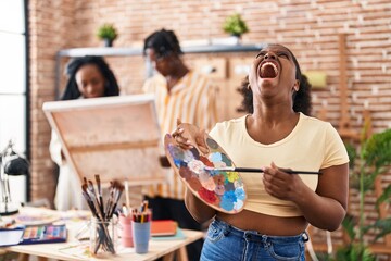 Young black painter woman at art studio holding palette angry and mad screaming frustrated and furious, shouting with anger looking up.