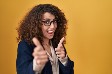 Hispanic woman with curly hair standing over yellow background pointing fingers to camera with happy and funny face. good energy and vibes.