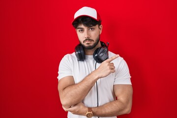 Hispanic man with beard wearing gamer hat and headphones pointing with hand finger to the side showing advertisement, serious and calm face