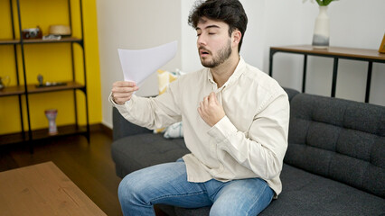 Young hispanic man sitting on sofa using paper as a hand fan at home