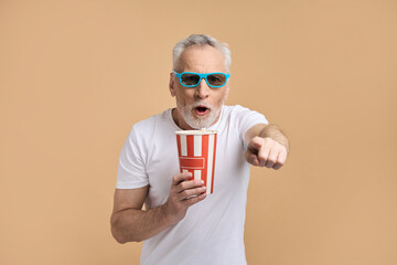 Excited senior man wearing 3d glasses holding popcorn bucket, watching movie pointing finger,  isolated on beige background. Hobby, positive lifestyle concept