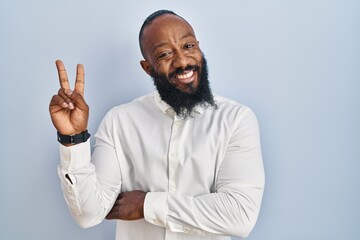 African american man standing over blue background smiling with happy face winking at the camera doing victory sign with fingers. number two.