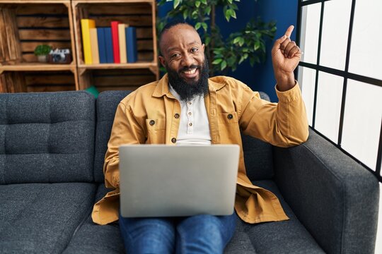 African american man using laptop at home sitting on the sofa smiling happy pointing with hand and finger to the side