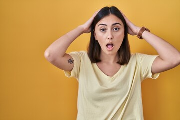 Hispanic girl wearing casual t shirt over yellow background crazy and scared with hands on head, afraid and surprised of shock with open mouth